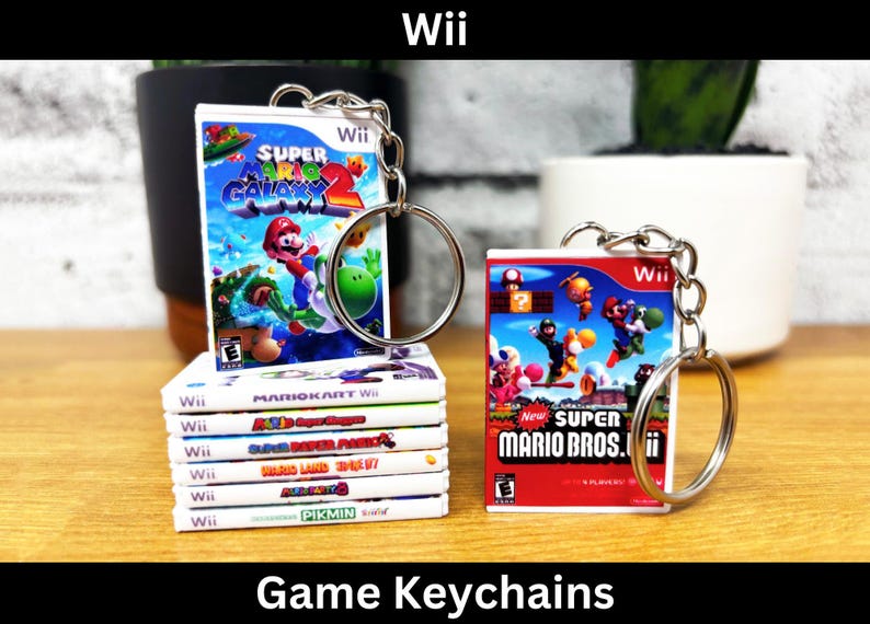 May include: Game keychains featuring miniature Wii game cases. One keychain shows "Super Mario Galaxy 2" and another "New Super Mario Bros. Wii". A stack of other Wii game cases is also visible. The word "Wii" is at the top of the image.