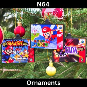 May include: A Christmas tree decorated with four ornaments shaped like Nintendo 64 game cartridges. The ornaments feature the games Super Smash Bros, Super Mario 64, The Legend of Zelda: Ocarina of Time, and a red and gold Christmas ornament.