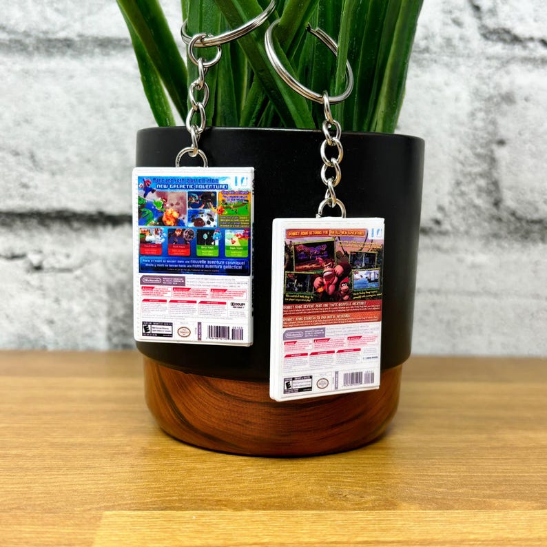 May include: Two keychains featuring miniature video game box art. One keychain displays a colorful game scene with the text "NEW GALACTIC ADVENTURE". The other shows a game scene with the text "DONKEY KONG". Both have silver key rings.