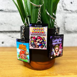 May include: Four miniature Nintendo GameCube game cases on keychains. The cases are for the games Paper Mario: The Thousand-Year Door, Animal Crossing, Luigi's Mansion, and a fourth game with a blurry title.