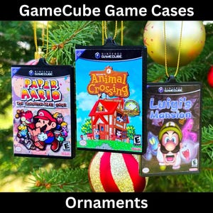May include: Three GameCube game cases are hanging from a Christmas tree as ornaments. The cases are for the games Paper Mario: The Thousand-Year Door, Animal Crossing, and Luigi's Mansion.