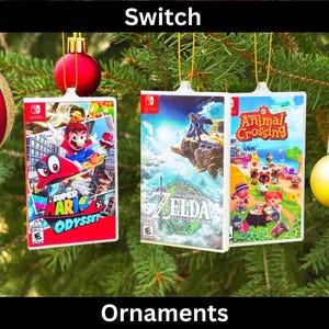 May include: Three Christmas ornaments shaped like Nintendo Switch game cases. The ornaments feature the game covers for Super Mario Odyssey, The Legend of Zelda: Breath of the Wild, and Animal Crossing: New Horizons. The ornaments are hanging on a green Christmas tree.
