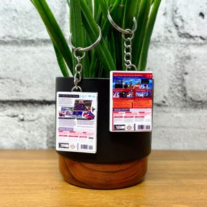 May include: Two keychains with miniature video game box art. The keychains are rectangular with silver key rings and chains. The game boxes feature colorful graphics and text. One keychain has the text "Get Behind the Wheel!" and the other has the text "More laps are on the adventure!"