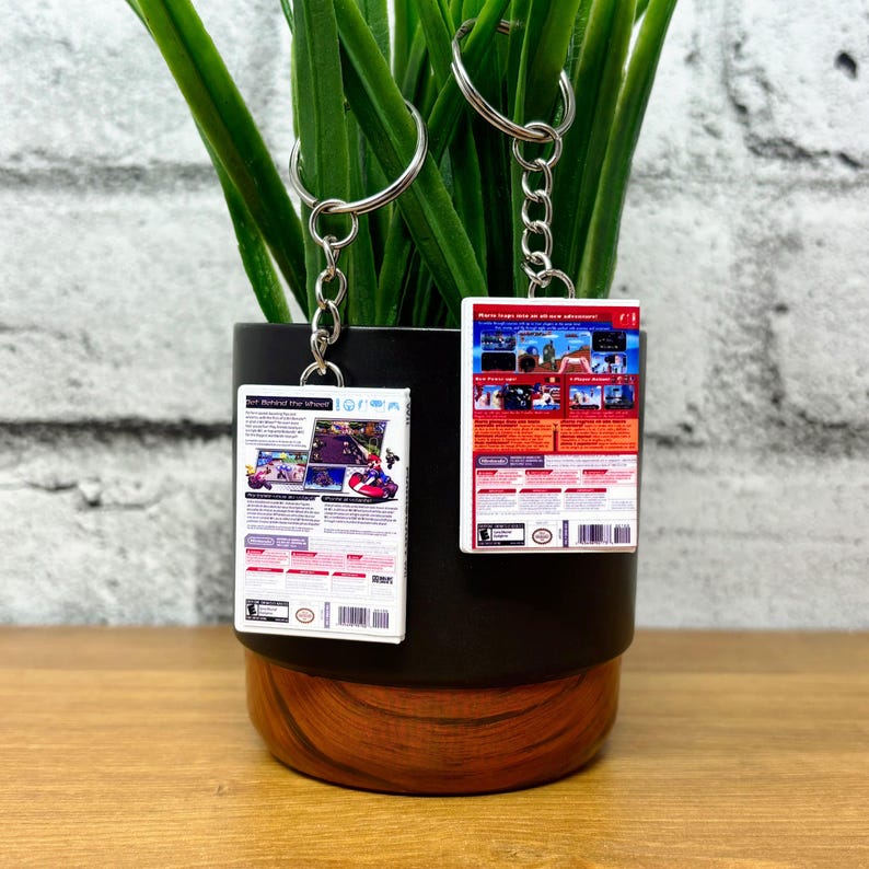 May include: Two keychains with miniature video game box art. The keychains are rectangular with silver key rings and chains. The game boxes feature colorful graphics and text. One keychain has the text "Get Behind the Wheel!" and the other has the text "More laps are on the adventure!"