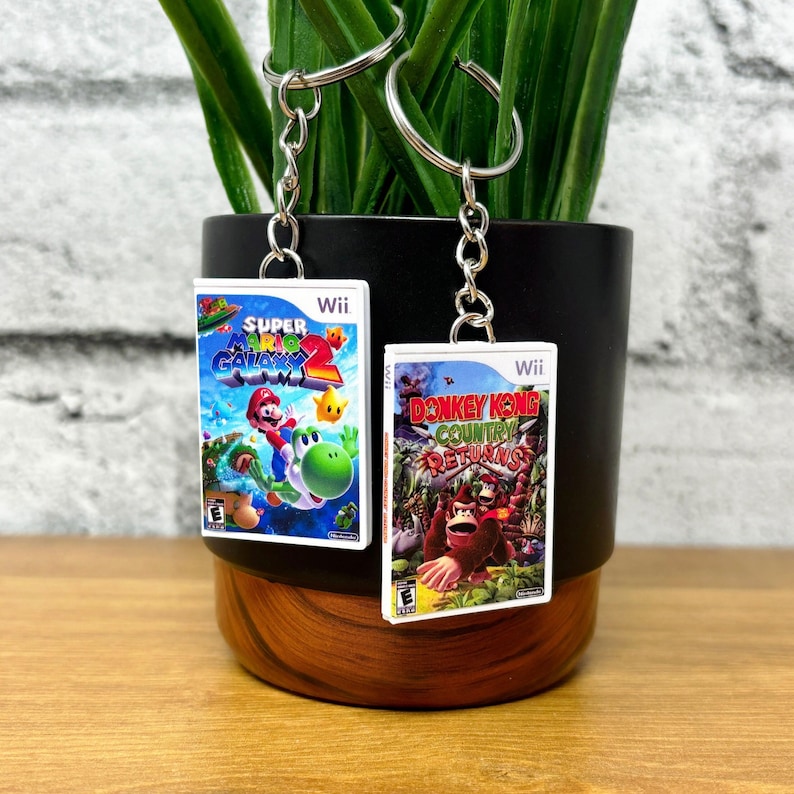May include: Two keychains featuring miniature game box art. One shows "Super Mario Galaxy 2" with Mario and Yoshi, the other "Donkey Kong Country Returns." Both have silver key rings and chains, and are set against a black and brown background.