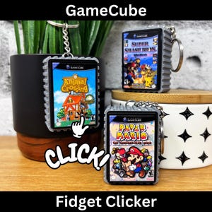Puede incluir: Llaveros GameCube Fidget Clicker con las portadas de los juegos Animal Crossing, Super Smash Bros y Paper Mario. Los llaveros tienen un marco gris texturizado y un llavero plateado. La palabra "CLICK" está en blanco, y el texto "Fidget Clicker" está debajo.