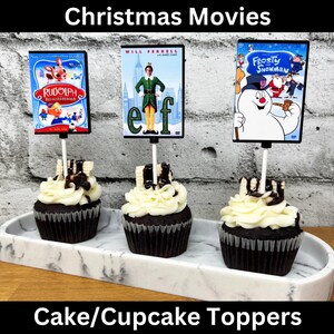 May include: Three chocolate cupcakes with white frosting and chocolate drizzle topped with movie themed cake toppers. The toppers are images of the films "Elf", "Rudolph the Red-Nosed Reindeer", and "Frosty the Snowman". The cupcakes are on a white marble cake stand. Cake/Cupcake Toppers