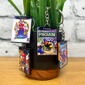 May include: Four miniature GameCube game cases on keyrings. The cases are for the games Super Mario Sunshine, Pikmin, Super Smash Bros. Melee, and Tony Hawk's Underground. The keyrings are hanging from a plant.