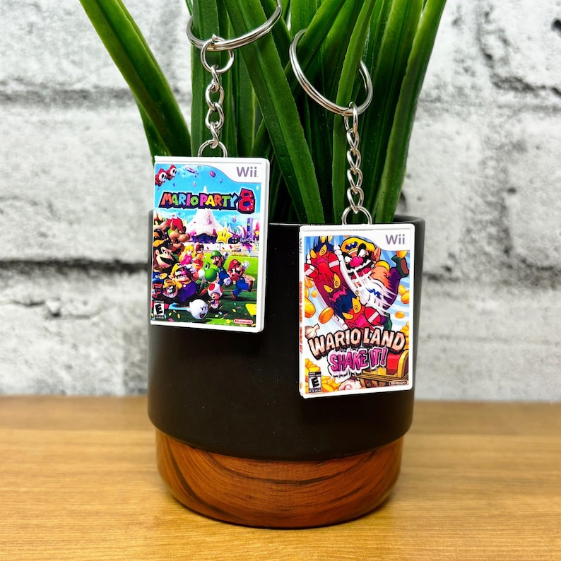 May include: Two keychains featuring miniature Wii game cases. One keychain displays "Mario Party 8" with colorful characters, and the other shows "Wario Land Shake It!" with a vibrant design. Both have silver key rings.
