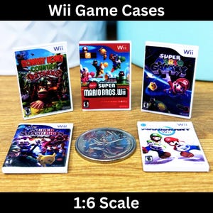 Op de afbeelding: Vijf miniatuur Wii-speldozen, elk met een ander spel. De dozen zijn wit met kleurrijke spelkunst en titels. De spellen zijn Donkey Kong Country Returns, Super Mario Bros. Wii, Super Smash Bros. Brawl, Super Mario Galaxy en Super Mario Galaxy 2. De dozen zijn in een rij op een houten oppervlak gerangschikt. De tekst "Wii" is zichtbaar op elke doos. De tekst "1:6 Scale" is zichtbaar onder de dozen.