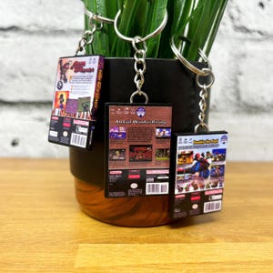 May include: Three miniature video game cases on keychains. The cases are black with colourful graphics and text. The text on the cases reads "Super Mario Strikers", "An Evil Wind is Rising", and "Double the Fun".
