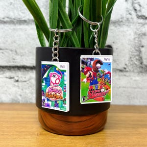 May include: Two keychains featuring miniature Wii game cases. One keychain displays "Kirby's Epic Yarn" and the other "Mario Super Sluggers." Both have silver key rings and chains. The keychains are displayed in front of a black and brown pot with green plants.
