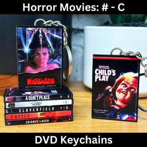 May include: A set of six horror movie DVD keychains. The keychains feature covers from popular horror movies such as "A Quiet Place", "Cloverfield", "Carrie", and "28 Days Later".  The keychains are black with red and white text.  One keychain features the movie "Child's Play" with a red and white title and a picture of Chucky.