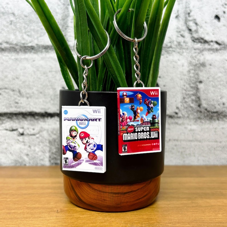 May include: Two keychains featuring miniature Wii game cases. One displays "Mario Kart Wii" with Mario and Luigi, the other "New Super Mario Bros. Wii." Both have silver key rings and chains, perfect for gamers.