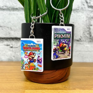 May include: Two keychains featuring miniature Wii game cases. One displays "Super Paper Mario" with Mario, Peach, and other characters. The other shows "Pikmin" with colorful creatures. Both have silver key rings and chains.