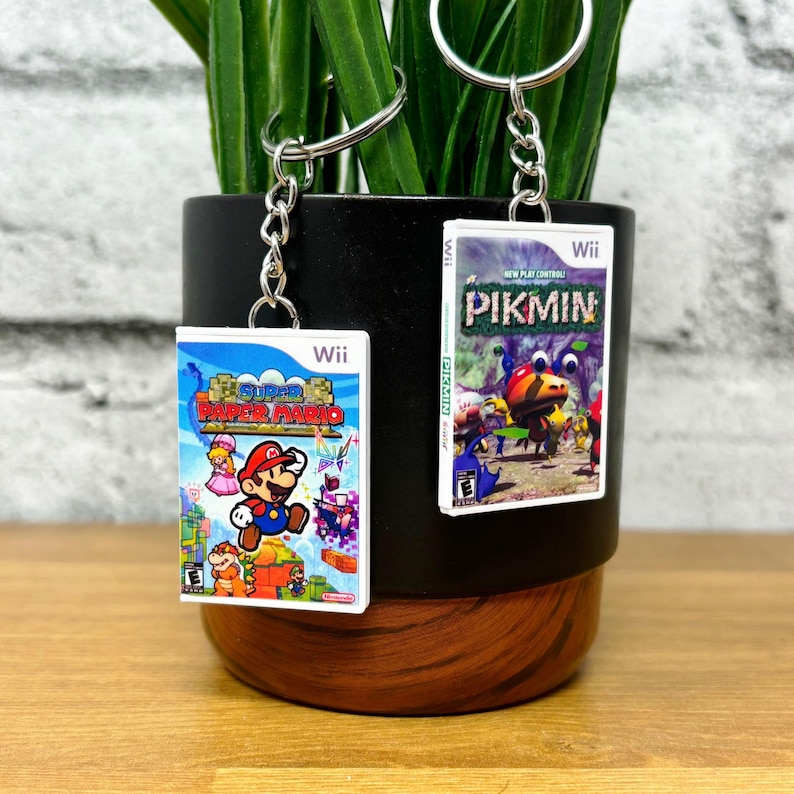 May include: Two keychains featuring miniature Wii game cases. One displays "Super Paper Mario" with Mario, Peach, and other characters. The other shows "Pikmin" with colorful creatures. Both have silver key rings and chains.