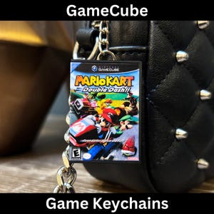 Puede incluir: Un llavero de GameCube con la carátula del juego Mario Kart: Double Dash!!. El llavero está sujeto a una cadena plateada y cuelga de un bolso negro con tachuelas. La imagen incluye el texto "GameCube" y "Game Keychains".