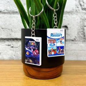 May include: Two keychains featuring miniature game box art. One displays "Super Mario Galaxy" with Mario, and the other shows "Wii Sports Resort." Both have silver key rings and chains, perfect for video game enthusiasts.