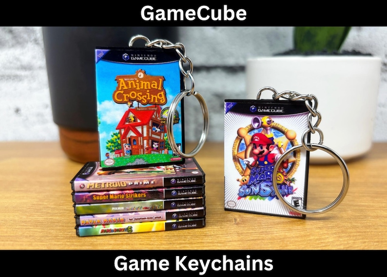 May include: Two GameCube game keychains. One keychain features the Animal Crossing game cover art with a red house and a blue sky. The other keychain features the Super Mario Sunshine game cover art with Mario in a blue and yellow suit. Both keychains are attached to silver key rings.