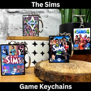 May include: Keychains featuring The Sims game covers. The keychains showcase various game versions, including The Sims, The Sims 2, and The Sims 4. Each keychain has a silver chain and a rectangular design with vibrant game artwork.
