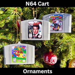 Puede incluir: Tres adornos grises de cartuchos de N64 colgados de un árbol de Navidad. Uno presenta a James Bond, otro a Mario Kart y el tercero a Super Mario 64. Se muestra el texto "N64 Cart" y "Ornaments".