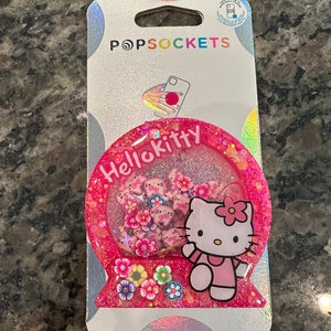 May include: A pink PopSockets phone grip featuring Hello Kitty. The grip has a glittery, snow globe-like design with Hello Kitty figures and flowers inside. The packaging is white with the PopSockets logo.