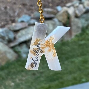 May include: A white resin letter K keychain with gold flakes embedded in the resin. The keychain has a gold chain and ring. The letter K has the name "Hailyn" written on it in black.