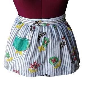 May include: Vintage half apron with a blue and white striped pattern. The apron features colorful floral and leaf appliqués in red, yellow, green, and black. A green pocket with white stitching is on the left side. The apron is displayed on a mannequin.