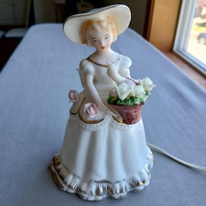 May include: A porcelain figurine of a woman in a white dress and hat, holding a basket of white roses. The figurine has gold trim and pink rose accents. A white electrical cord extends from the base.