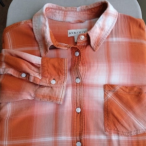 May include: Orange and white plaid button-down shirt with a chest pocket and buttoned cuffs. The shirt features the brand name "AVA VIV" on a tag near the collar. The fabric has a faded, textured appearance.