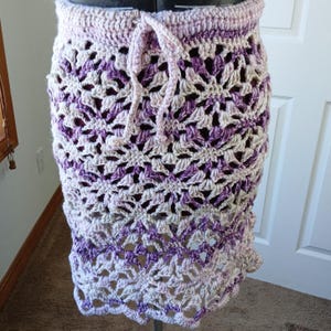 May include: A crocheted skirt with a purple and white ombre pattern. The skirt has a drawstring waist and a scalloped hem.