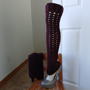 May include: A pair of dark burgundy crochet leg warmers, one worn over a silver glitter high heel shoe. The leg warmer has an open, lacy design. A matching crochet cuff sits beside the shoe. The items are displayed on a wooden chair.