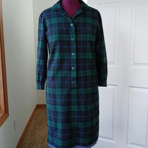 May include: A knee-length dress featuring a dark green, navy blue, and black plaid pattern. The garment has long sleeves, a collared neckline, and a front button-down closure with five matching buttons. The fabric has a textured appearance.