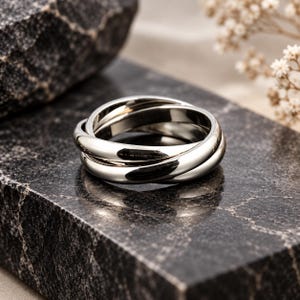 Op de afbeelding: Een gepolijste zilverkleurige ring bestaande uit drie in elkaar grijpende banden die elkaar overlappen om een doorlopende cirkelvorm te creëren. Het metalen oppervlak is glad en reflecterend met een heldere afwerking en subtiele accenten.