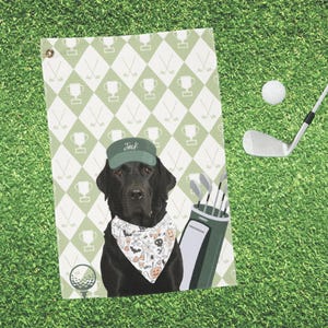 May include: Golf towel with a green and white diamond pattern, golf clubs, and a black Labrador wearing a green cap with the name "Jack" and a Halloween bandana. A golf ball and club are on the green grass.