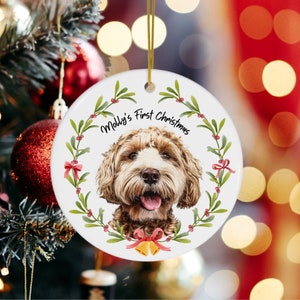 Op de afbeelding: Een witte kerstbal van keramiek met het gezicht van een bruine en witte hond in het midden. De kerstbal is versierd met een groene krans met rode bessen en een rode strik. De tekst "Molly's First Christmas" staat in het zwart op de kerstbal.