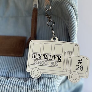 May include: A white wooden bus rider tag with black text that reads "Bus Rider School Bus #28". The tag is attached to a blue corduroy backpack with a brown leather strap.