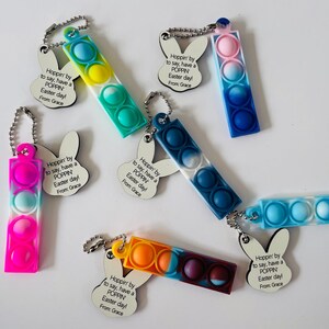 May include: A set of six colorful pop-it fidget toys with bunny-shaped keychains. Each keychain has a tag that reads "Hoppin' by to say, have a POPPIN Easter day! From Grace".