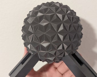 Könnte beinhalten: Eine dunkelgraue, geometrische Kugel mit strukturierter Oberfläche, getragen von zwei abgewinkelten, rechteckigen Beinen. Die Kugel besteht aus zahlreichen dreieckigen Facetten und erzeugt ein einzigartiges, architektonisches Design. Das Objekt wird in einer Hand gehalten.
