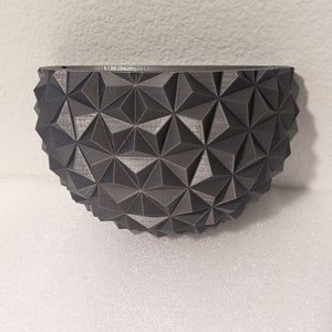 Könnte beinhalten: Eine dunkelgraue Schüssel mit geometrischem Muster und Halbkugelform. Die Oberfläche der Schüssel ist mit einem sich wiederholenden Muster aus dreieckigen Formen bedeckt, wodurch ein strukturiertes Aussehen entsteht. Die Schüssel besteht wahrscheinlich aus einem harten Material wie Kunststoff oder Keramik.