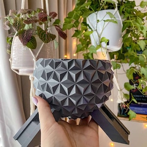 May include: A gray geometric planter with a textured surface, held in a hand. The planter has a spherical shape with a pattern of triangular facets. It is supported by three angled legs. Other hanging planters with plants are in the background.
