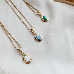 May include: Three gold-colored necklaces with oval pendants. The pendants feature gemstones in white, blue, and green. The necklaces are displayed on a cream-colored fabric, showcasing the jewelry's design and color variations.