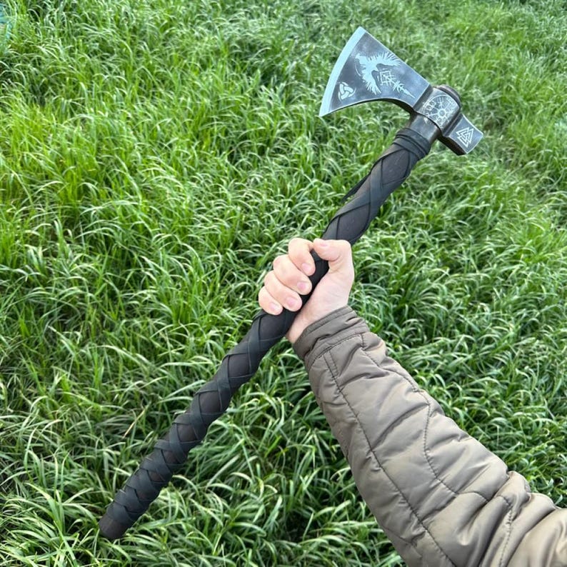 Op de afbeelding: Een bijl met een zwarte steel en een zilveren blad. Het blad heeft een Viking-ontwerp met een vogel en een symbool. De bijl wordt vastgehouden door een hand in een bruin jack.