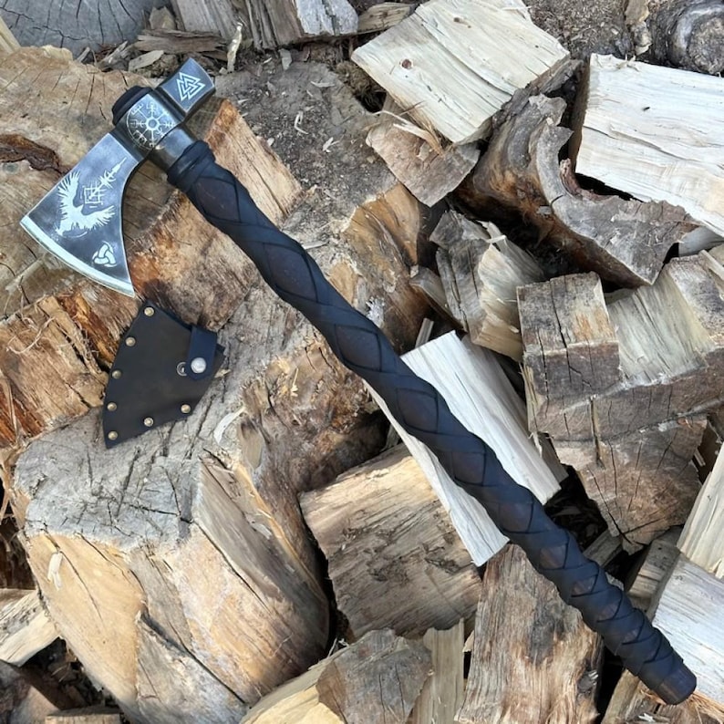 Op de afbeelding: Een bijl met een zwarte steel en een zilveren blad met een raaf en een triskel symbool. De bijl ligt op een stapel houtblokken.