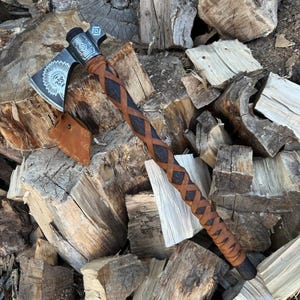 Könnte beinhalten: Eine handgeschmiedete Axt mit einem lederumwickelten Stiel. Der Axtkopf zeigt ein kunstvolles Design mit einem indianischen Kopfschmuck. Die Axt liegt auf einem Stapel Brennholz.