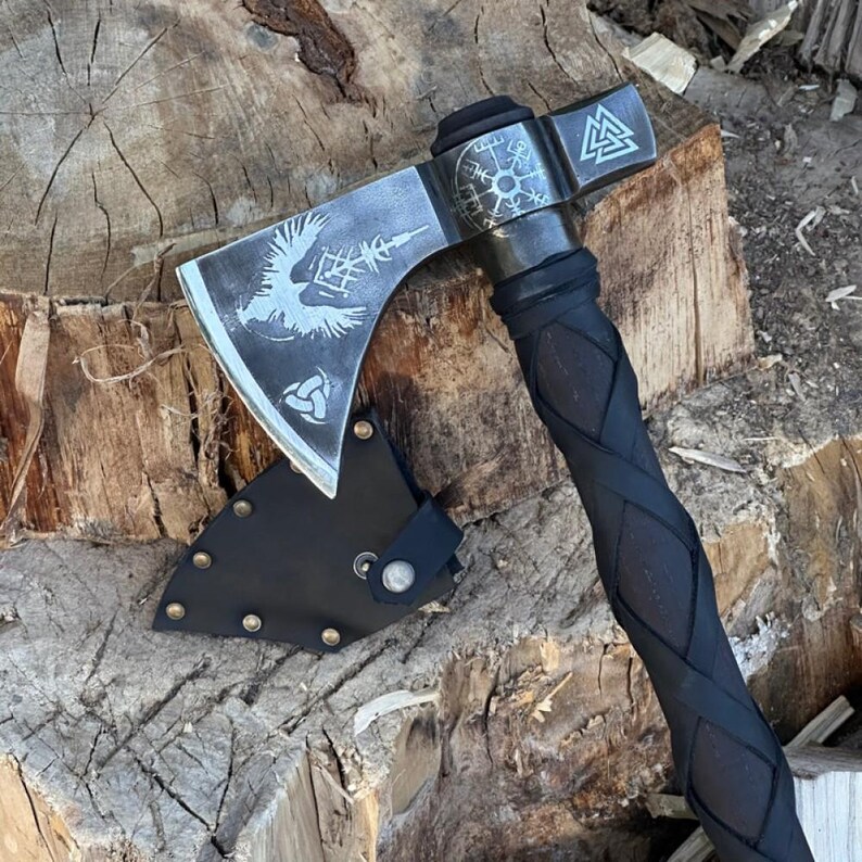 Op de afbeelding: Een zilveren bijl met een zwarte steel en een zilveren ontwerp van een vogel en een kompas op de kling. De bijl zit in een zwarte leren schede en ligt op een houten oppervlak.