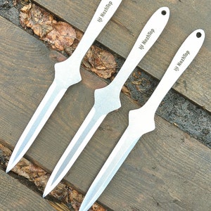 Puede incluir: Tres cuchillos plateados para lanzar con el texto "NozhTop" impreso en la hoja. Los cuchillos tienen una punta afilada y un mango en forma de diamante. Los cuchillos están dispuestos en diagonal sobre una superficie de madera.