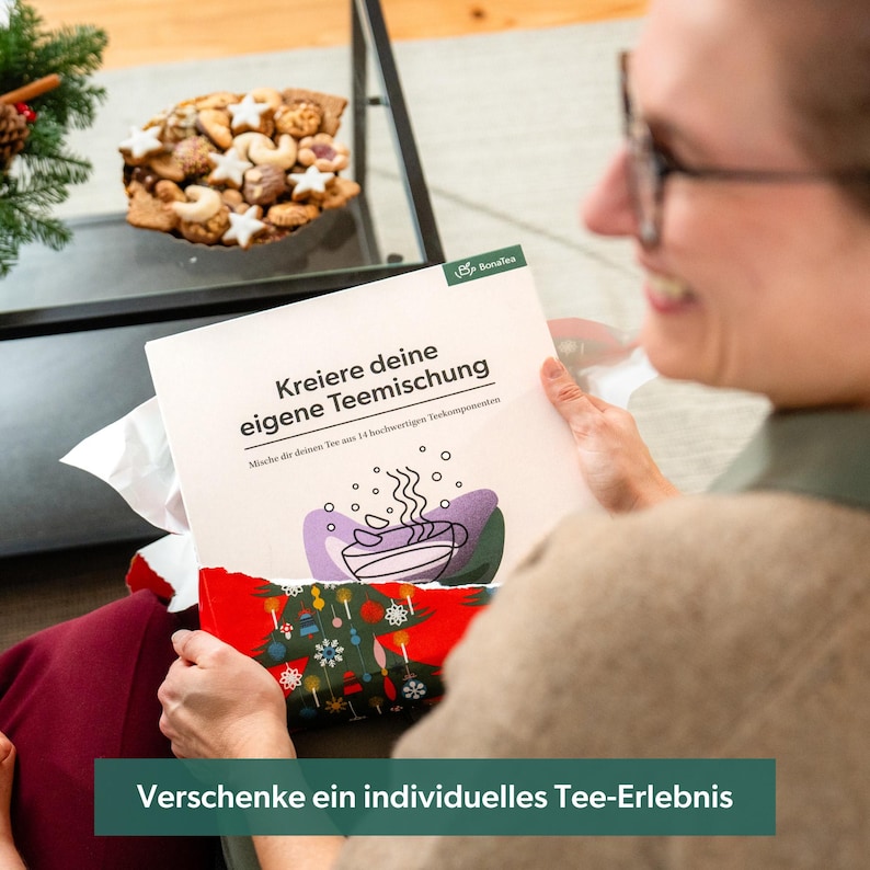 May include: A white booklet with the text "Kreiere deine eigene Teemischung" and a colorful illustration of a steaming cup of tea. The booklet is being held by a person's hands. The text "Verschenke ein individuelles Tee-Erlebnis" is at the bottom of the image.