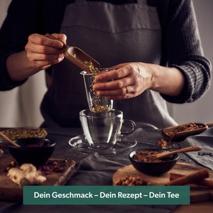 May include: A person is adding loose leaf tea to a glass tea infuser. The infuser is sitting on top of a glass teacup. There are various tea ingredients and a wooden spoon on the table. The text on the table reads "Dein Geschmack - Dein Rezept - Dein Tee".