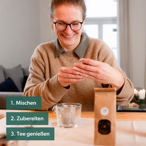 May include: A woman smiles as she holds a brown tea bag package. Three green rectangular boxes with white text are on the table: "1. Mix", "2. Prepare", and "3. Enjoy Tea".
